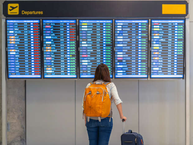 A woman stands with her back to the camera, holding the handle of a navy suitcase. She is looking up at a blurry airport departure board and wearing a bright orange rucksack.